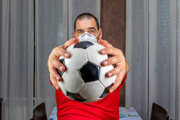 Sakallı, kısa saçlı, kırmızı tişörtlü ve maskeli bir adam bir futbol topuyla koronavirüsü önlemek için. Sağlıklı yaşam konsepti - Fotoğraf, Görsel