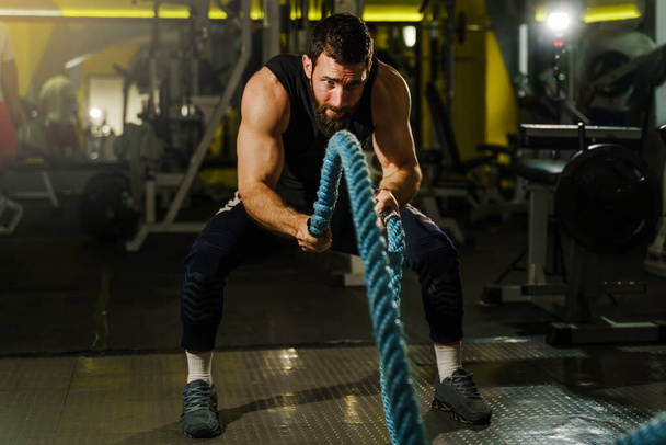 Fronz view portrait of young adult caucasian man male athlete training at the dark gym working out using battle rope wearing black shirt and beard strength and endurance training - Photo, Image