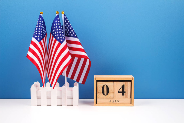 Sada tří amerických vlajek s datem 4. července na dřevěných cihlách na bílém stole s modrým pozadím. Národní symbol USA - vlajka Stará sláva. Koncept oslav Dne nezávislosti - Fotografie, Obrázek
