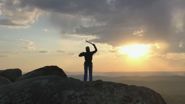 Slow motion drone filmato di un giovane felice arrampicatore maschio in piedi sulla cima di una montagna con successo alzando le braccia e tenendo una piccozza ascia e godendo di un momento al tramonto. - Filmati, video