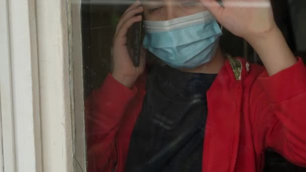 Ένα παιδί με ιατρική μάσκα κοιτάει από το παράθυρο και μιλάει στο τηλέφωνο. Αυτοαπομόνωση σε καραντίνα, coronavirus, covid19. - Πλάνα, βίντεο