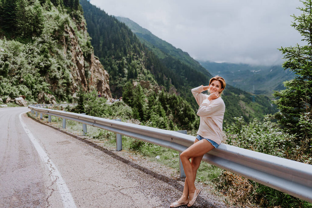 Young female traveler sitting near road enjoying mountain woods. A young woman enjoys the view of the mountain landscape, on one of the most famous roads in Europe, Transfagarasan, in the Fagaras Mountains in Romania, Europe. Mountain woodland landsc - Photo, Image