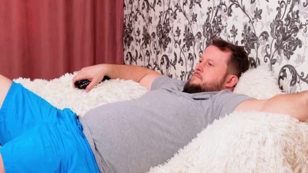 髭を生やしたハンサムな太った男はチャンネルを切り替えるためにリモコンを使ってテレビを見ている。男はテレビの画面で見たものにうんざりしている。自宅のリビングルームのソファに横になり、スペースをコピーします. - 映像、動画