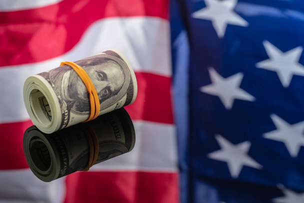 Ρολό των χρημάτων, αμερικανικά δολάρια 100 χαρτονομίσματα σε φόντο καθρέφτη με αμερικανική σημαία αντανακλάται σε αυτό - Φωτογραφία, εικόνα