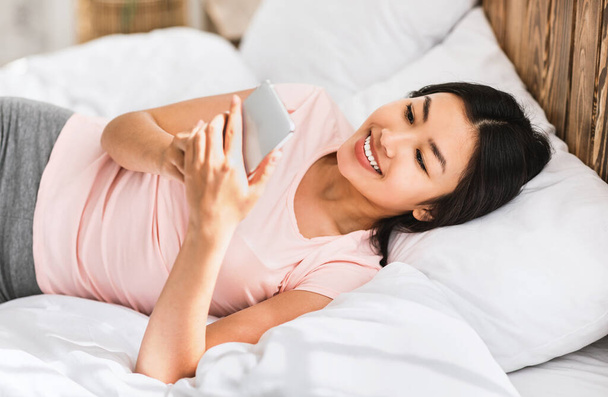 Азіатська дівчина зі смартфоном використовуючи мобільний додаток бреше в ліжку
 - Фото, зображення
