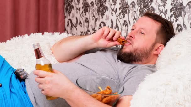 Grubas pije piwo i je niezdrowe skrzydełka kurczaka, znudzony przed telewizorem na kanapie. Koncepcja niedożywienia, kwarantanny w domu, alkoholizm. - Materiał filmowy, wideo