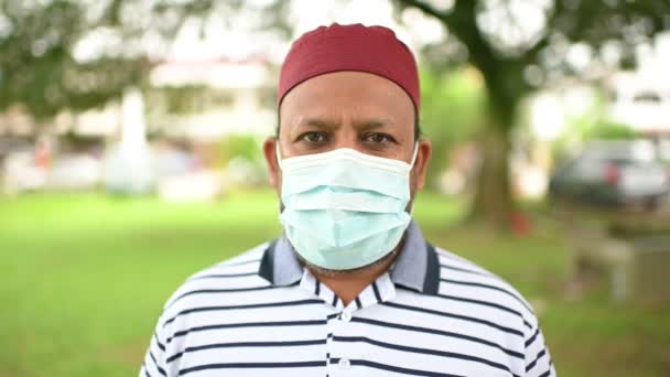 Ein asiatischer muslimischer Mann trägt eine Gesichtsmaske mit muslimischer Gebetsmütze und blickt in die Kamera, um sich vor einer Krankheit zu schützen, die heute weit verbreitet ist. Gesundheitswesen und medizinische Versorgung nach Covid-19-Konzept. - Filmmaterial, Video