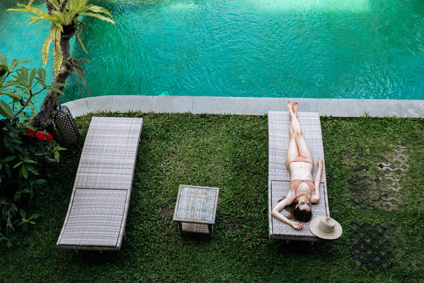 Top view of slim woman in beige bikini relaxing lying on lounge chair at the poolside near luxury pool in tropic on vacation - Fotó, kép