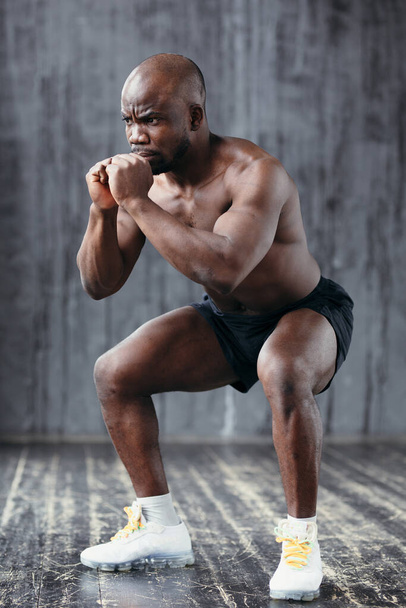 Athletic bare-chested African American doing squats while holding hands near chest in gym - Foto, Imagem