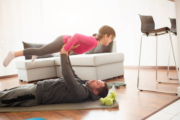 Ojciec i córka trenują w domu. Ćwiczenia w mieszkaniu. Sport w domu. Tatuś leży na macie do jogi i trzyma córkę w ramionach. - Zdjęcie, obraz