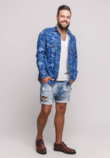 Stylish young cheerful guy with a beautiful beard in a denim shirt and denim shorts on a simple gray background. Denim shirt and denim shorts advertising concept for billboard - Fotó, kép