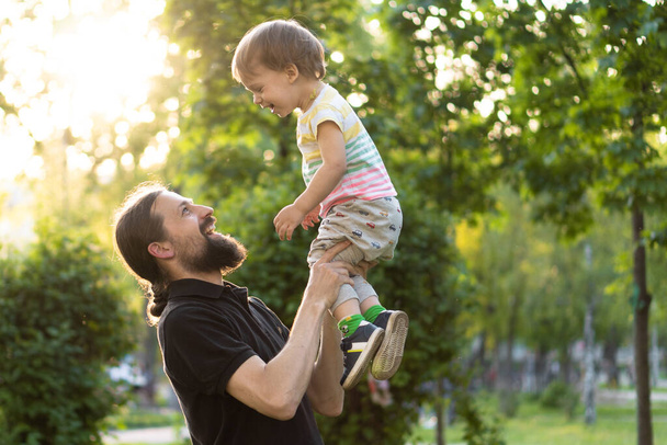 Fatherhood, parenthood, childhood, caring, summer and leisure concept - young dad with beard and long hair in black t-shirt holds in his arms little son in the backlight of the sunset in the park. - Zdjęcie, obraz