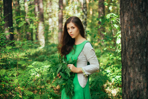 A young, attractive woman in a green dress from a medieval villager holds a fern leaf in her hands. A village herbalist collects herbs for a potion. - Foto, afbeelding