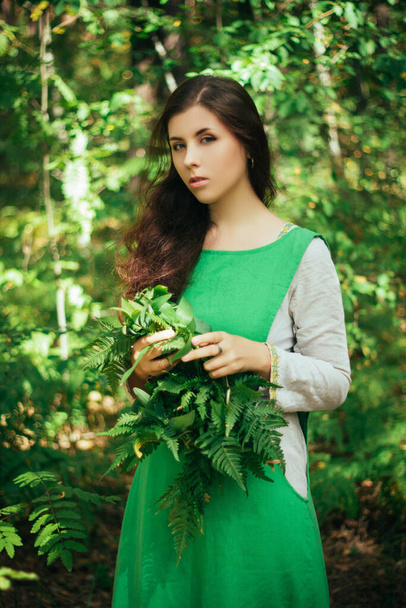 A young, attractive woman in a green dress from a medieval villager holds a fern leaf in her hands. A village herbalist collects herbs for a potion. - Фото, зображення