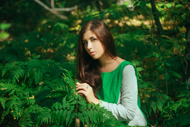 Attractive girl in a green medieval dress sits in the bushes of fern. Village woman herbalist collects herbs for potion in the forest. - Fotó, kép
