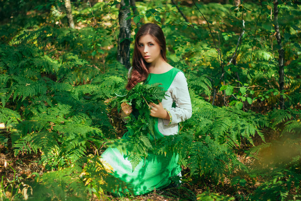 Attractive girl in a green medieval dress sits in the bushes of fern. Village woman herbalist collects herbs for potion in the forest. - Фото, изображение