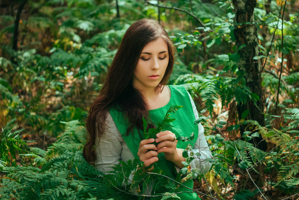 Viehättävä tyttö vihreässä keskiaikaisessa mekossa istuu saniaisen pensaissa. Kylä nainen yrtti kerää yrttejä juoma metsässä
. - Valokuva, kuva