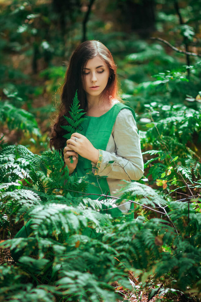 Muchacha atractiva en un vestido medieval verde se sienta en los arbustos de helecho. Mujer de pueblo herbolario recoge hierbas para poción en el bosque
. - Foto, Imagen