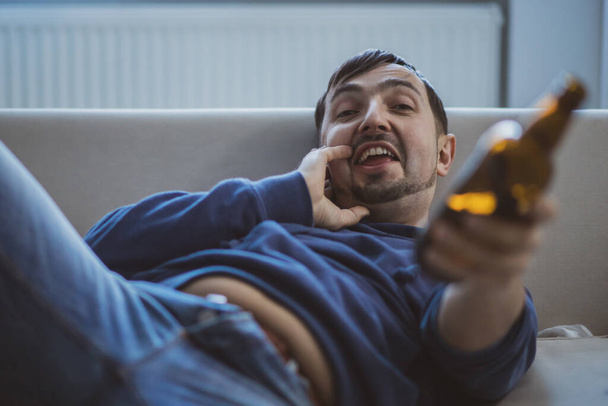 A man laying on a couch proposes a bottle of beer to drink. - Valokuva, kuva