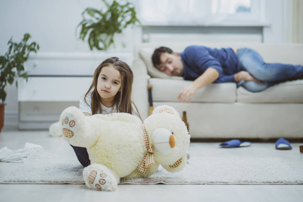 Sad daughter sitting with teddy bear while drunk father sleeping on sofa. - Photo, Image