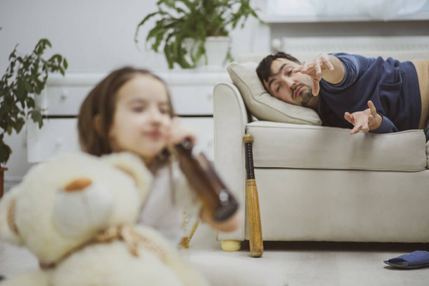 Little daughter trying to sip a beer while her dad is laying on a couch. - 写真・画像