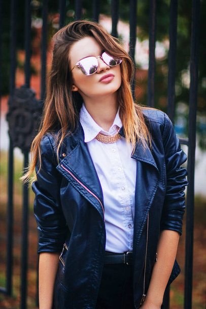 Young beautiful fashionable European woman in a black leather jacket on a background of a dark metal fence. - Photo, image