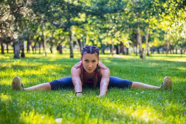 足を伸ばすための運動をするスポーツ女性は、スプリットに座っています。芝生の上の公園で. - 写真・画像