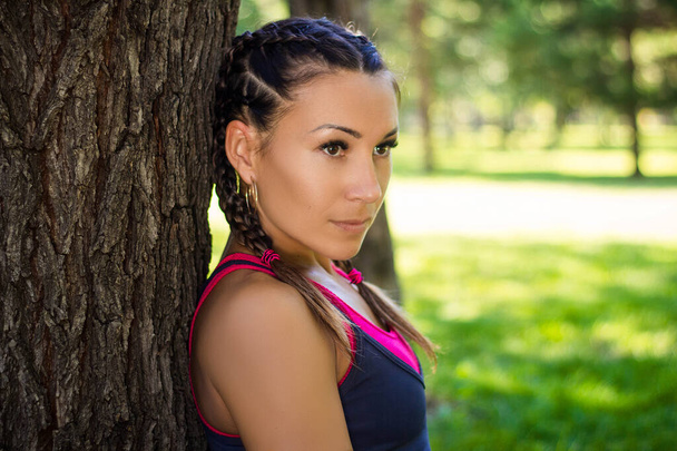 Athletic woman standing on the grass leaning against a tree in the park on the background of trees - Фото, зображення