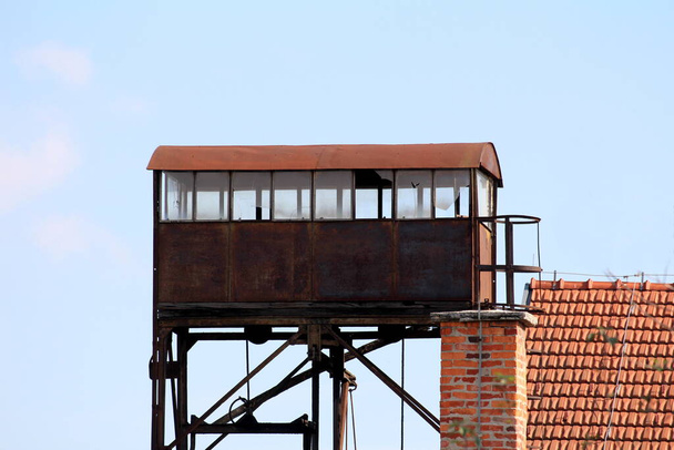 Rusted old vintage metal abandoned railway station observation tower with broken windows mounted on strong metal construction next to red bricks building on clear blue sky background - Photo, Image