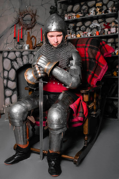 A young knight in medieval armor with a sword in his hand sits in a rocking chair by the fireplace. - Foto, afbeelding
