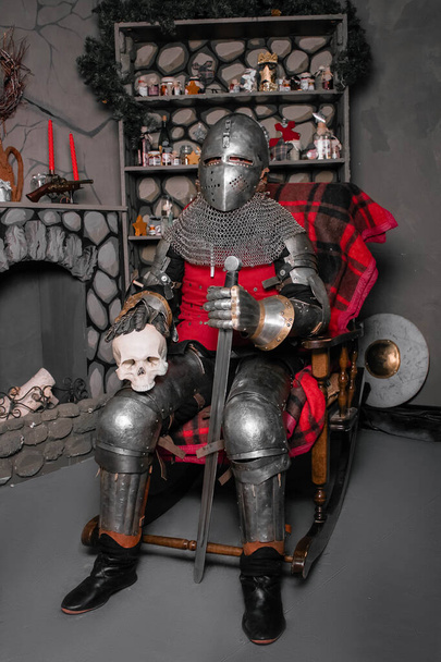 Jovem cavaleiro de armadura do século XIV com uma espada numa mão e um crânio na outra mão, sentado numa cadeira de balanço junto à lareira
. - Foto, Imagem