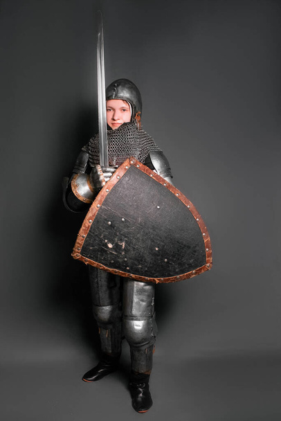 Un joven caballero con armadura medieval armado con una espada y un escudo de pie sobre un fondo gris
. - Foto, Imagen