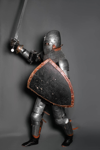 A medieval knight in armor with a shield and a sword in his hands stands in a fighting position and waves his weapon. - Foto, Imagem