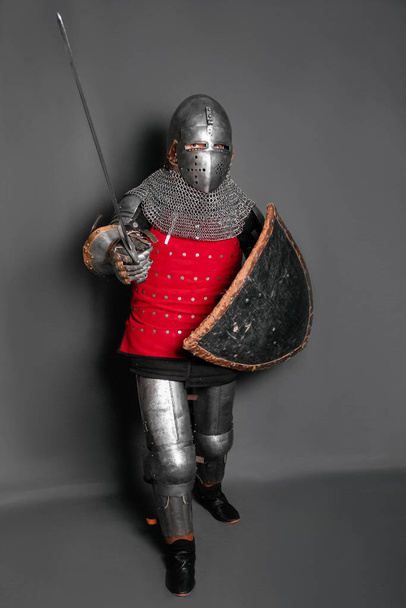 A medieval knight in armor with a shield and a sword in his hands stands in a fighting position and waves his weapon. - Photo, image
