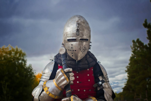 Knight in helmet and red brigantine against the background of thunderclouds. With a two-handed sword in his hands. - Foto, imagen