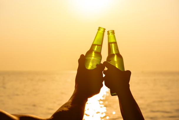Deux mains tenant des bouteilles de bière serrant et célébrant les vacances à la plage au coucher du soleil d'été
 - Photo, image