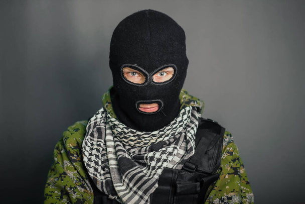 A man in camouflage and bullet proof vest, with a balaclava on his head. A special unit soldier, a modern warrior. A photo on a gray background, studio. - Fotografie, Obrázek