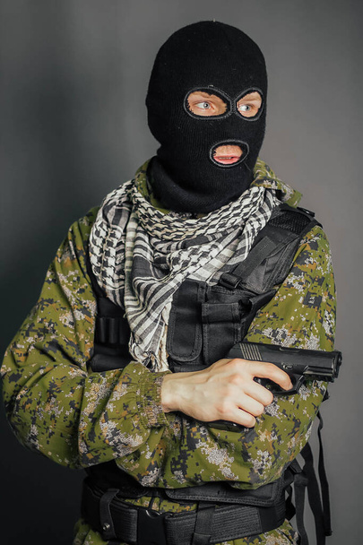 Portrait of a soldier in camouflage uniform, body armor and balaclava holding a gun in his hands, standing on a gray background. - Fotografie, Obrázek