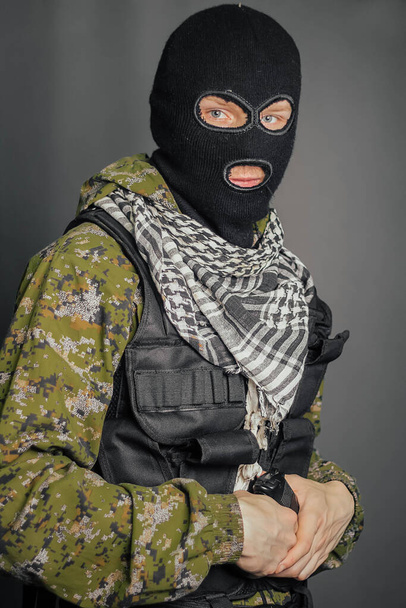 Portrait of a soldier in camouflage uniform, body armor and balaclava holding a gun in his hands, standing on a gray background. - Фото, изображение