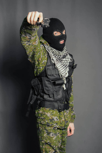 A man in camouflage uniform, body armor and a balaclava, holds his weapon ready and takes aim with a pistol, standing against a gray background. - Foto, Bild