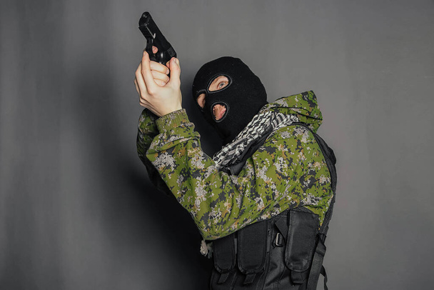 A man in camouflage uniform, body armor and a balaclava, holds his weapon ready and takes aim with a pistol, standing against a gray background. - Photo, image