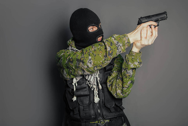 Ein Mann in Tarnuniform, Panzer und Sturmhaube hält seine Waffe bereit und zielt mit einer Pistole auf einen grauen Hintergrund. - Foto, Bild