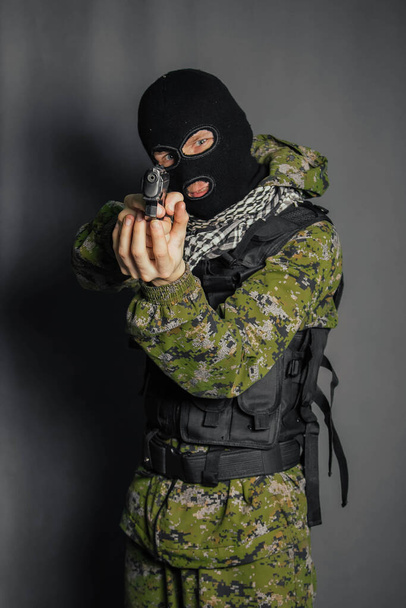 A man in camouflage uniform, body armor and a balaclava, holds his weapon ready and takes aim with a pistol, standing against a gray background. - Foto, immagini