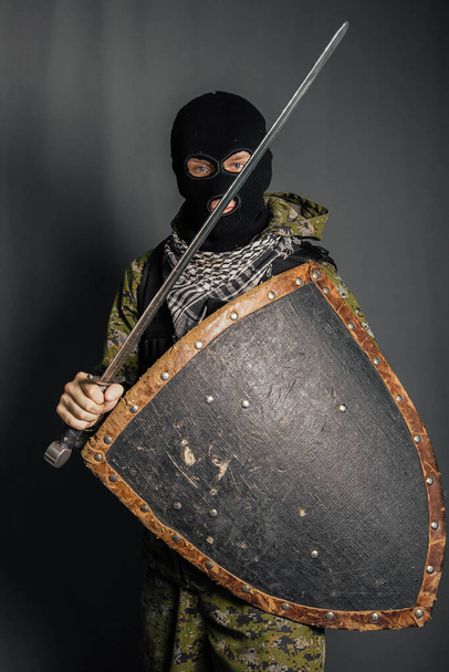 Σύγχρονος πολεμιστής με μεσαιωνικά όπλα στα χέρια του. Ένας στρατιώτης υπερασπίζεται τον εαυτό του με μια ασπίδα και επιτίθεται με ένα σπαθί. Προστασία του νόμου, του νόμου, της τάξης, της ιδιοκτησίας, του χρήματος, της ειρήνης του μυαλού. - Φωτογραφία, εικόνα