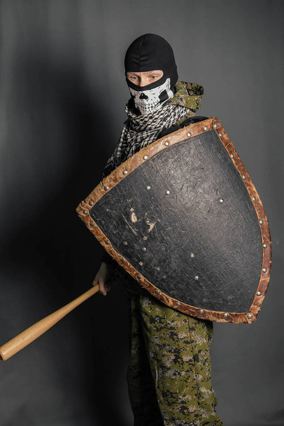 A radical protesting man in camouflage uniform with a balaclava in the form of a skull on his head holds a shield and a baseball bat in his hands. Calls for battle. Studio photo on a gray background. - Photo, Image