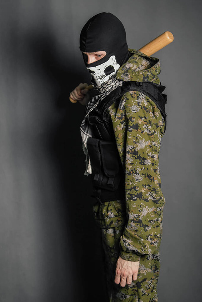 Unknown man, a robber in a balaclava with a skull, in camouflage uniform and body armor with a baseball bat in his hands. Studio photo on a gray background. - Fotoğraf, Görsel