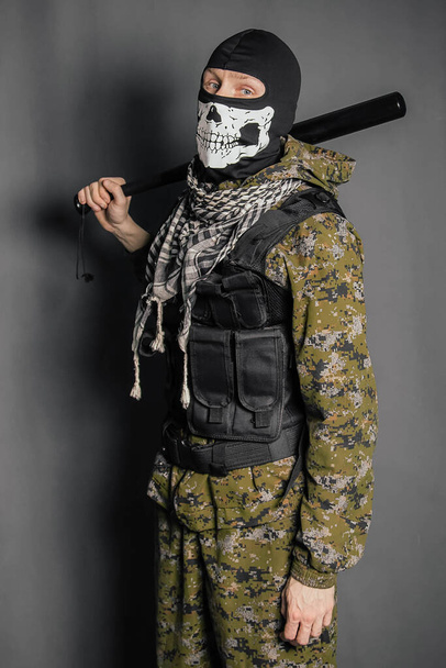 Unknown man, a robber in a balaclava with a skull, in camouflage uniform and body armor with a baseball bat in his hands. Studio photo on a gray background. - Foto, imagen