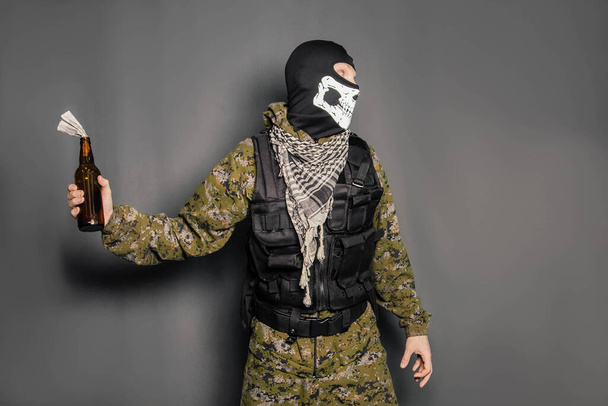 A man in a balaclava with a skull pattern, dressed in camouflage with a hood and a bulletproof vest, is holding a Molotov cocktail in his hands. A protester throws a Molotov cocktail. Studio photo on a gray background. - Photo, Image