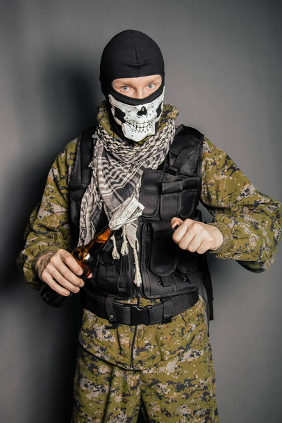A man in a balaclava with a skull pattern, dressed in camouflage with a hood and a bulletproof vest, is holding a Molotov cocktail in his hands. A protester throws a Molotov cocktail. Studio photo on a gray background. - Foto, Imagen
