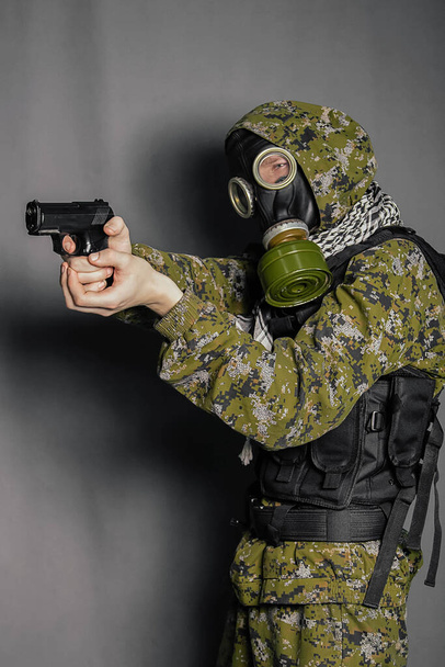 A soldier in camouflage uniform, body armor, in a gas mask with a hood on his head, holds a gun in his hands. Cosplay man, stalker. Studio photo on a gray background. - Photo, image